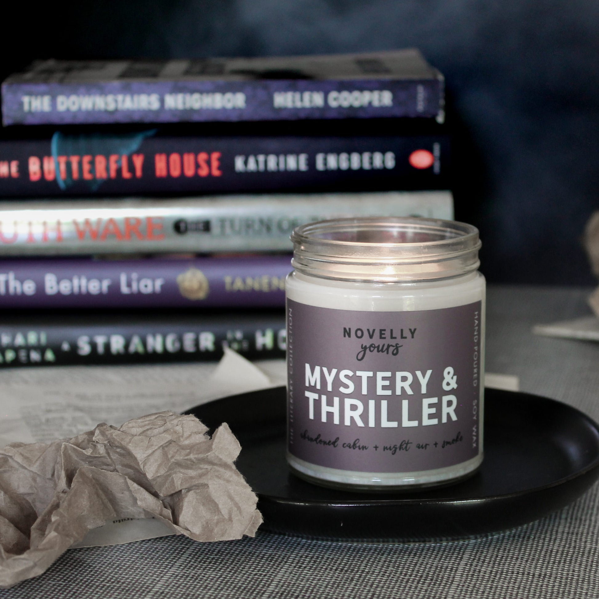 mystery & thriller scented soy wax candle with purple grey label and candle is lit. sits on black round tray with paper accents with thriller books in background