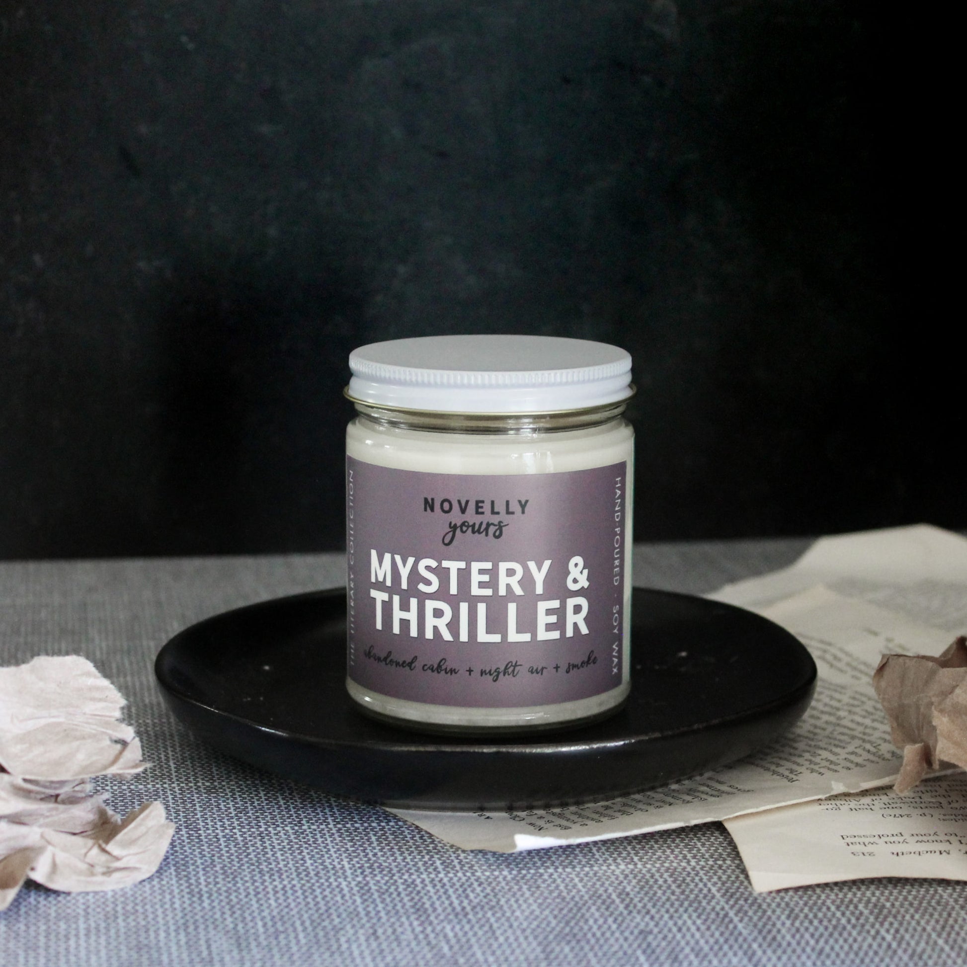 mystery & thriller scented soy wax candle with purple grey label and white lid sits on black round tray with paper accents