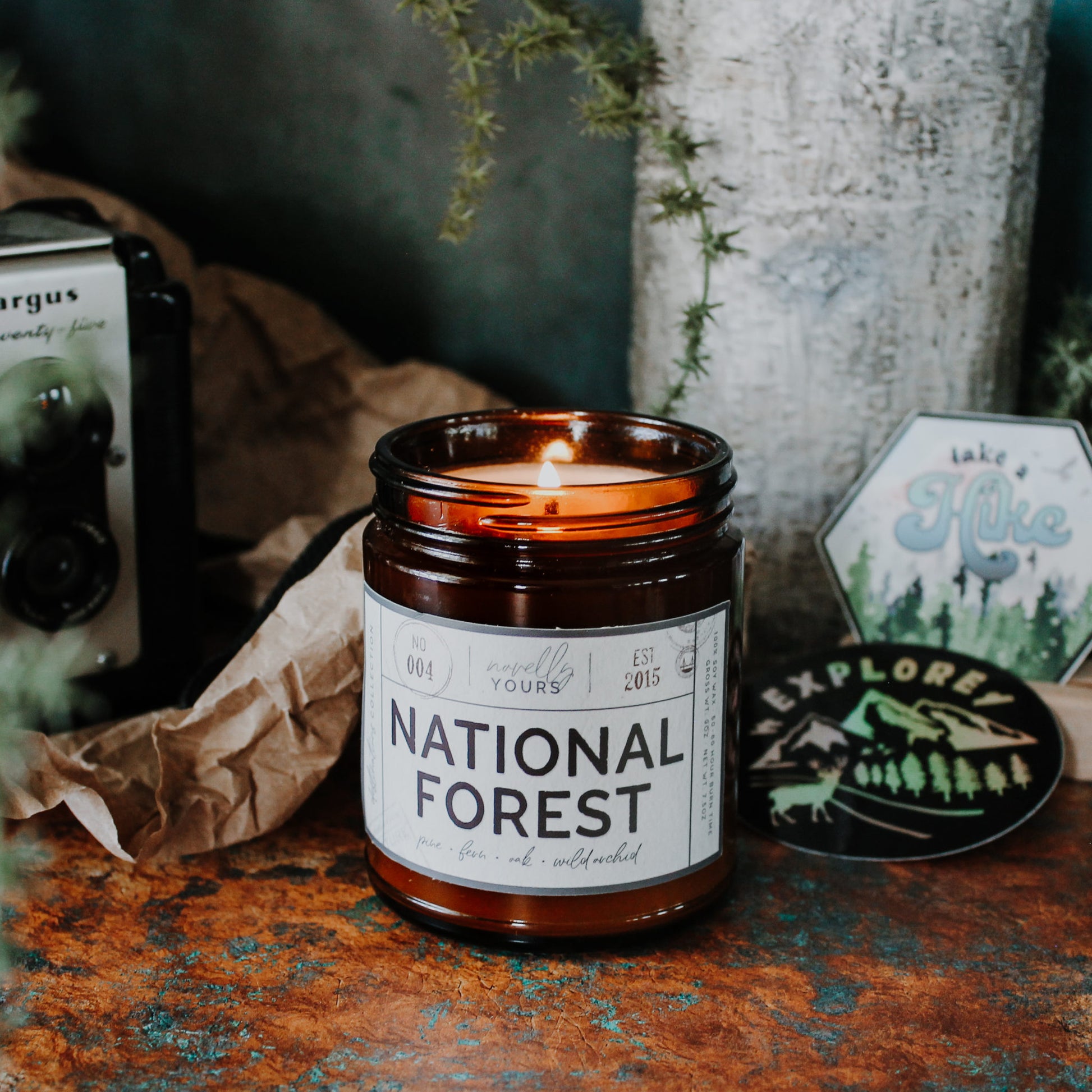 national forest scented soy wax candle in amber jar. candle is lit, sits on rustic surface surrounded by relevant decor
