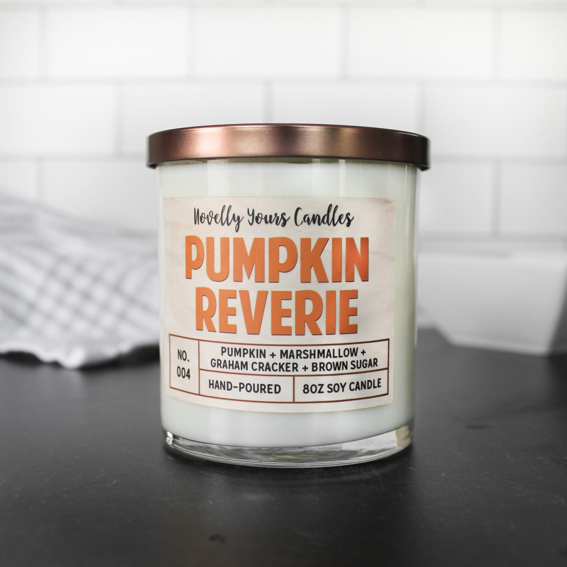 pumpkin reverie fall scented candle, pumpkin marshmallow scented candle. Candle is in clear tumbler glass jar with bronze lid. Sits on black surface with white subway tile background.