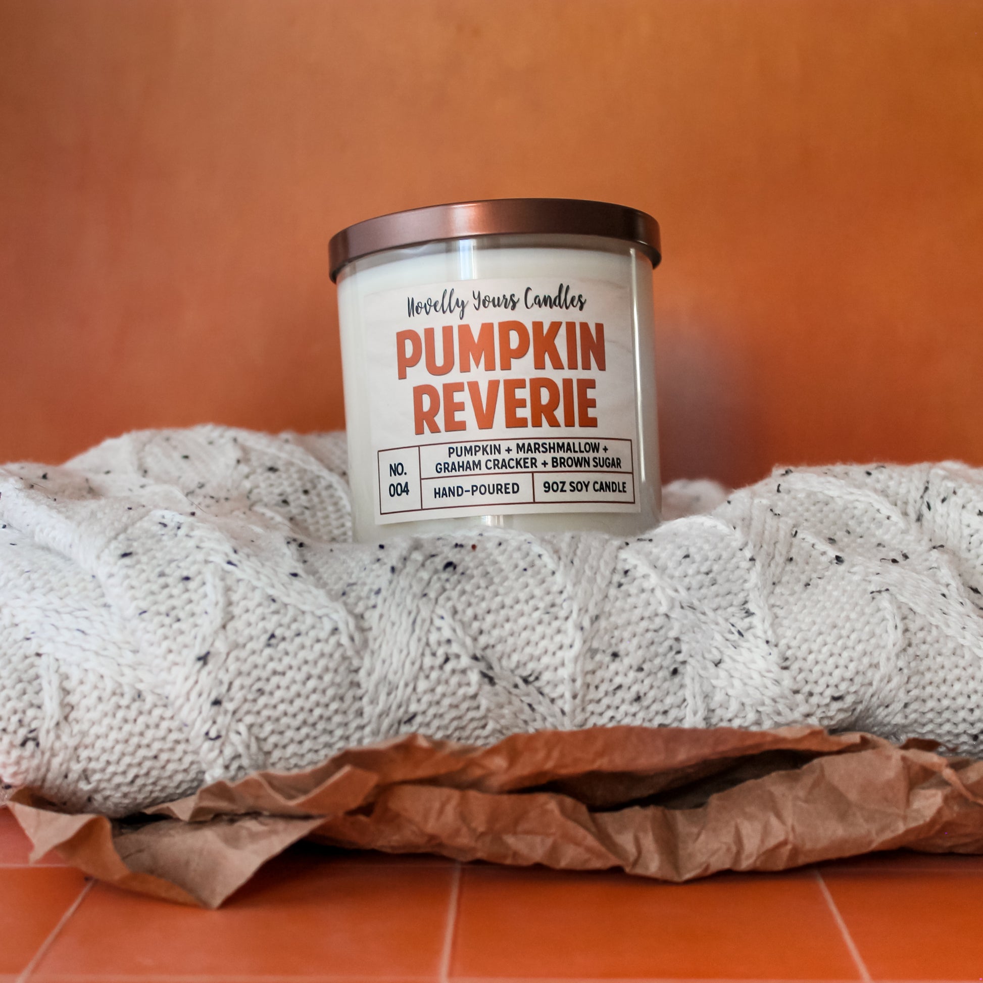 pumpkin reverie fall scented candle, pumpkin marshmallow scented candle. Candle is in clear tumbler glass jar with pumpkin colored text. Candle sits sweater and Kraft paper with orange background, all on top of orange tile counter