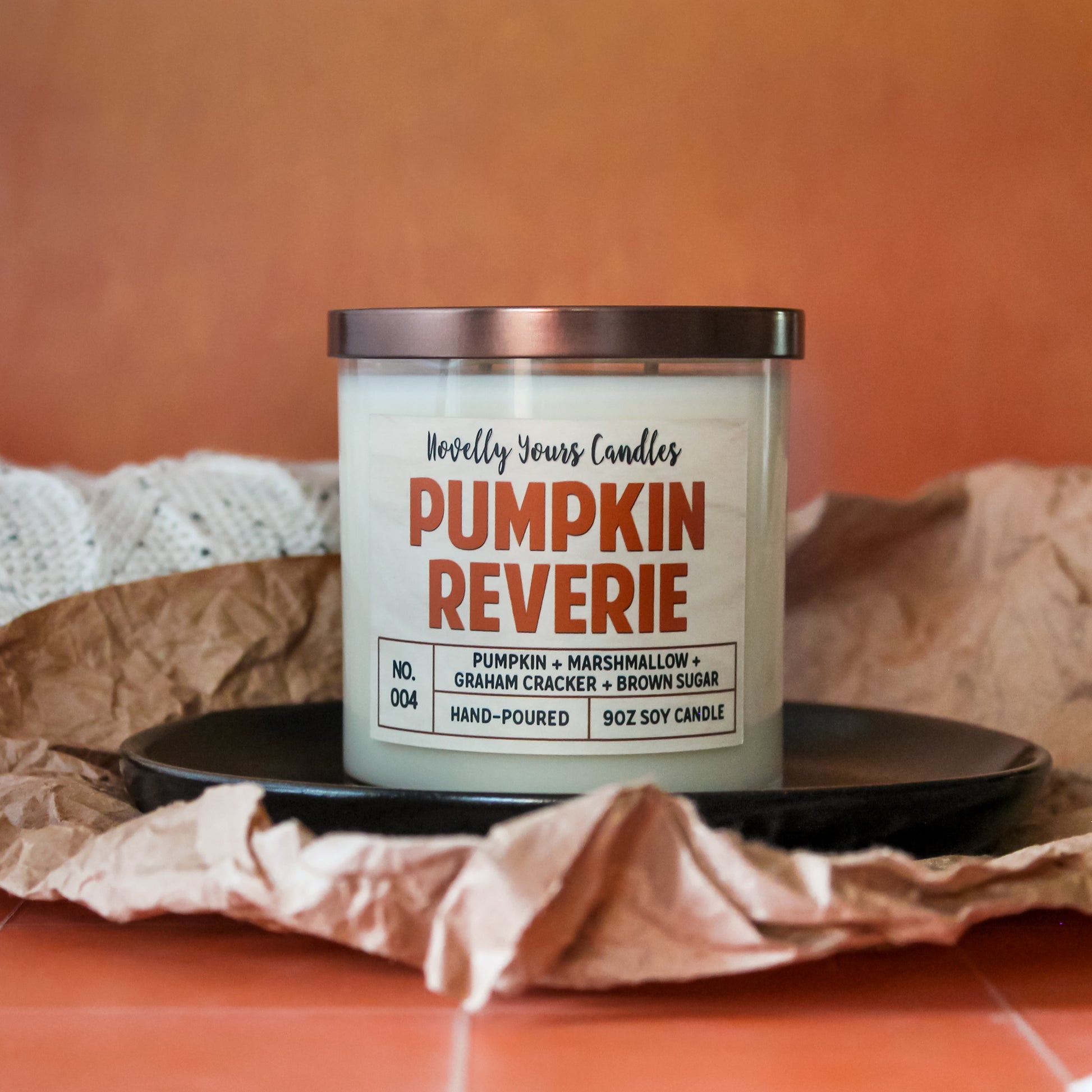 pumpkin reverie fall scented candle, pumpkin marshmallow scented candle. Candle is in clear tumbler glass jar with pumpkin colored text. Candle sits on black plate on orange tile counter, surrounded by sweater and Kraft paper with orange background
