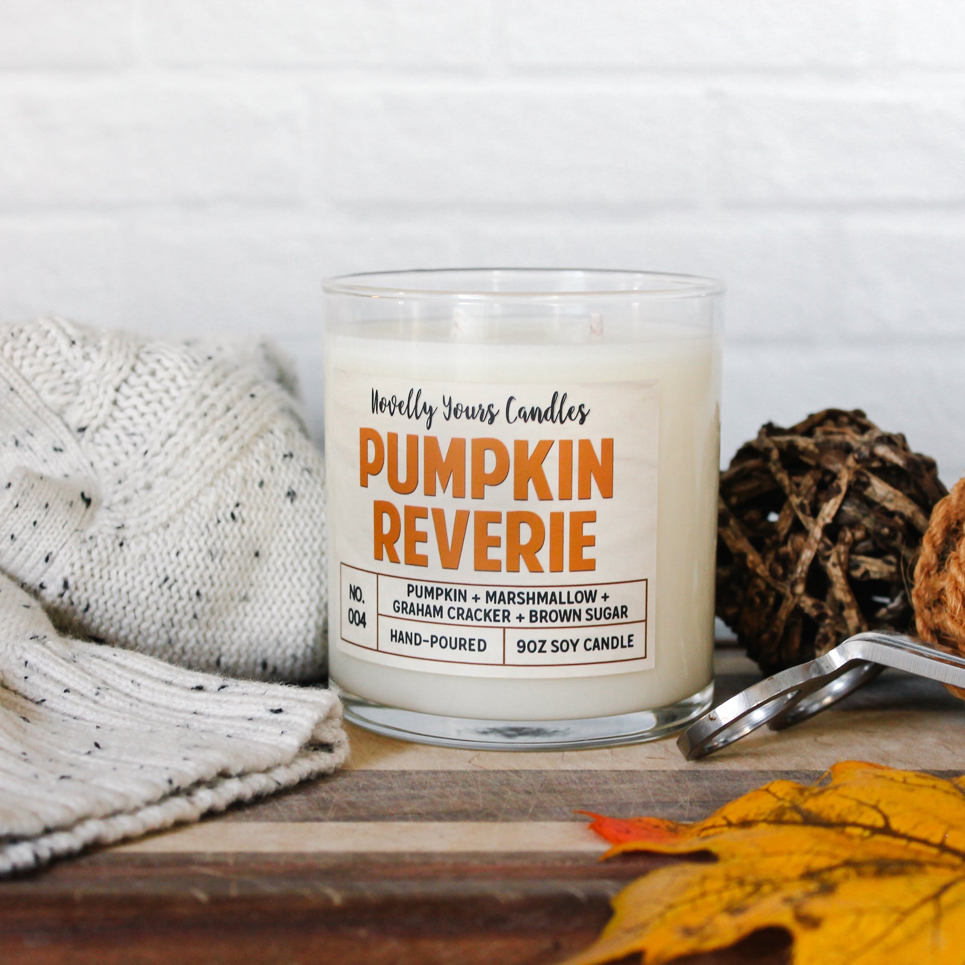 pumpkin reverie fall scented candle, pumpkin marshmallow scented candle. Candle is in clear tumbler glass jar with pumpkin colored text. Candle sits on wooden cutting board surrounded by autumn decor