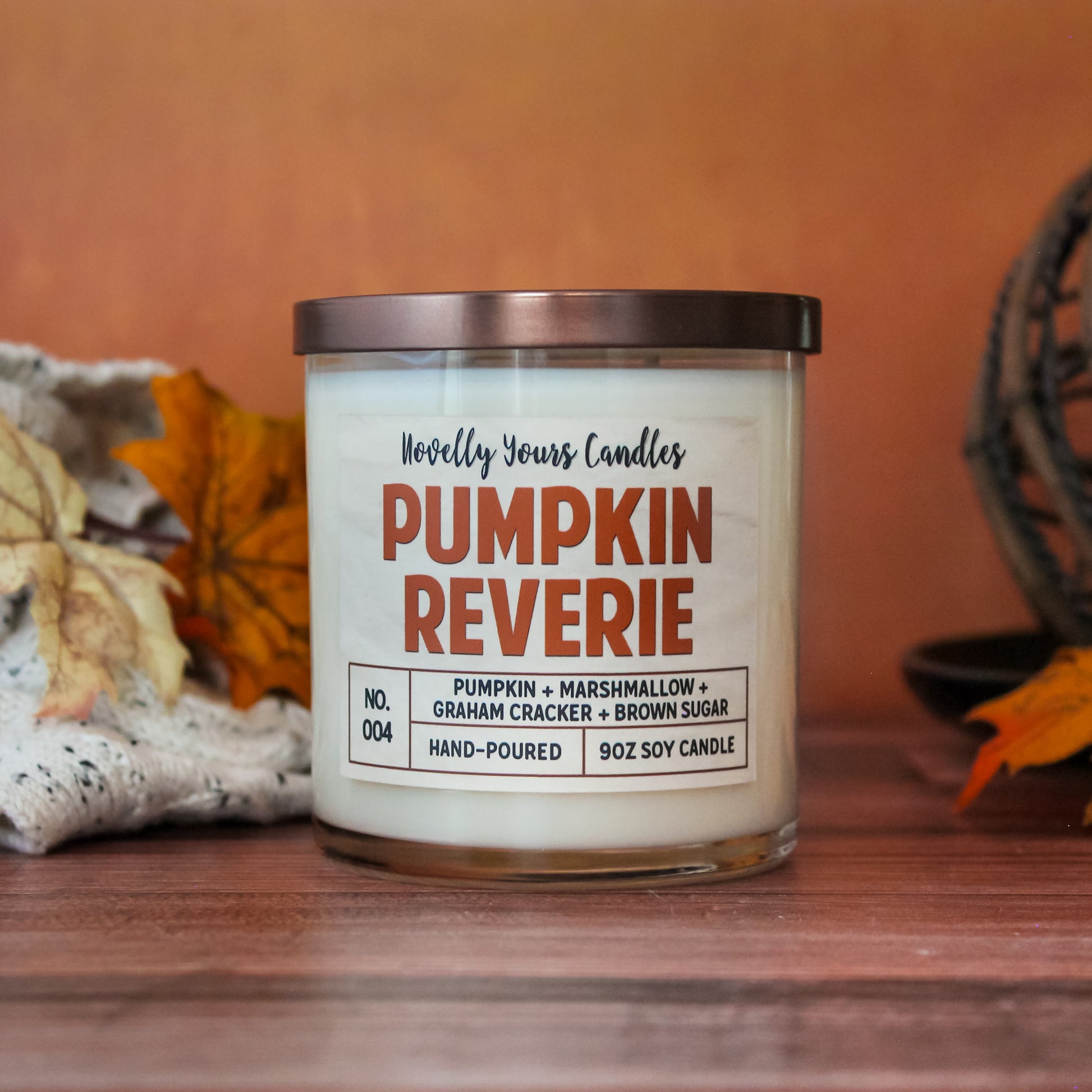 pumpkin reverie fall scented candle, pumpkin marshmallow scented candle. Candle is in clear tumbler glass jar with pumpkin colored text. Candle sits on wooden surface with orange background, surrounded by autumn decor