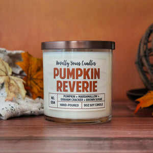 pumpkin reverie fall scented candle, pumpkin marshmallow scented candle. Candle is in clear tumbler glass jar with pumpkin colored text. Candle sits on wooden surface with orange background, surrounded by autumn decor