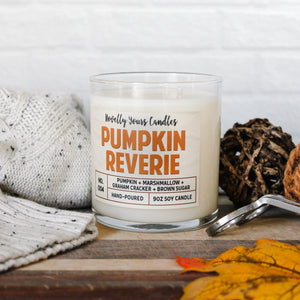 pumpkin reverie fall scented candle, pumpkin marshmallow scented candle. Candle is in clear tumbler glass jar with pumpkin colored text. Candle sits on wooden cutting board surrounded by autumn decor