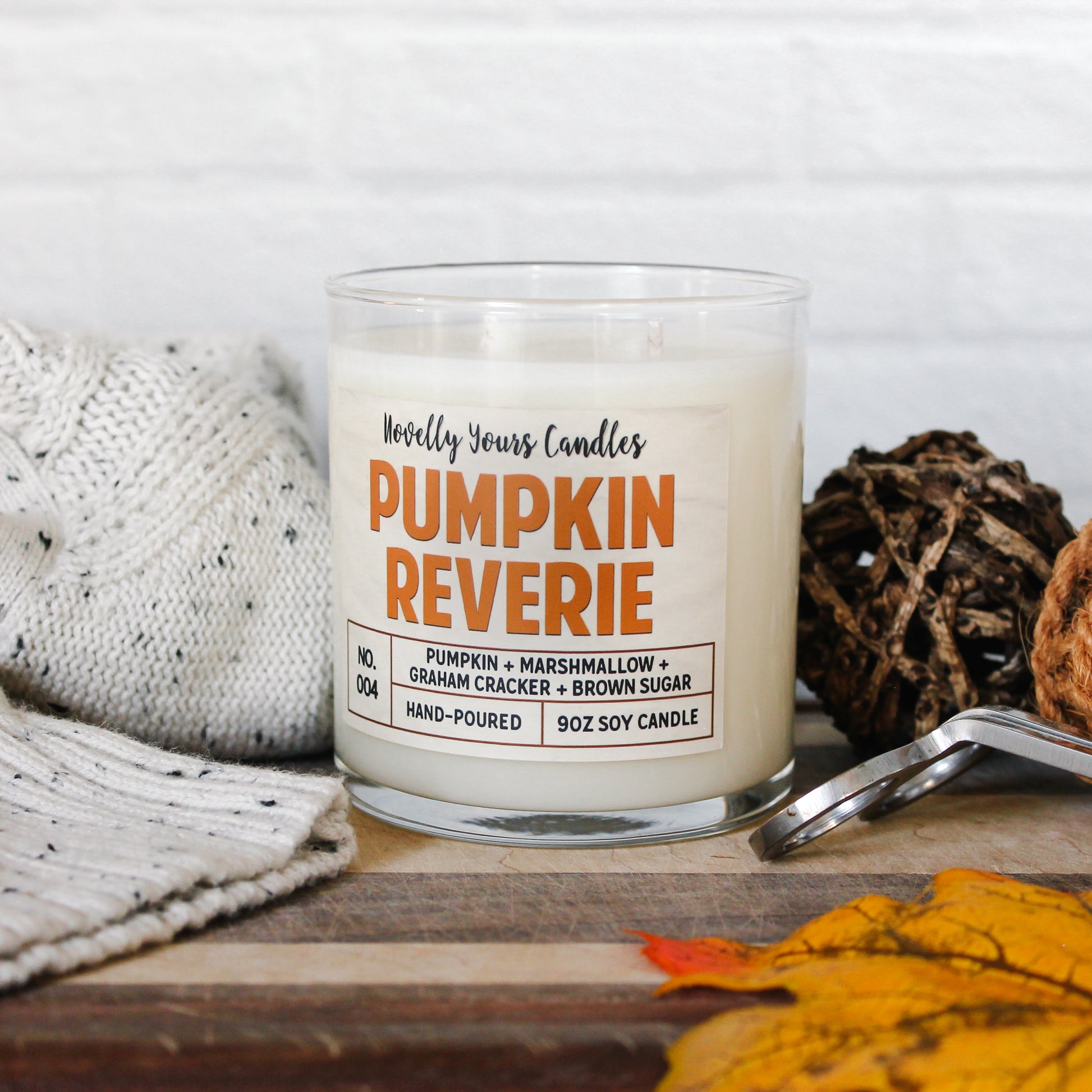 pumpkin reverie fall scented candle, pumpkin marshmallow scented candle. Candle is in clear tumbler glass jar with pumpkin colored text. Candle sits on wooden cutting board surrounded by autumn decor