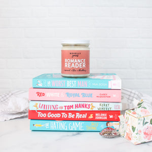 romance reader scented soy wax candle in clear glass jar with white lid and soft rose colored label on top of pink and blue book stack of romance books