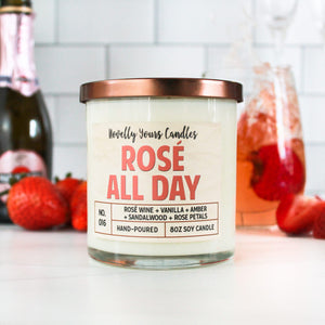 Rosé All Day scented soy wax candle in glass tumbler jar with bronze lid and pink label lettering surrounded by rose' wine and fruit
