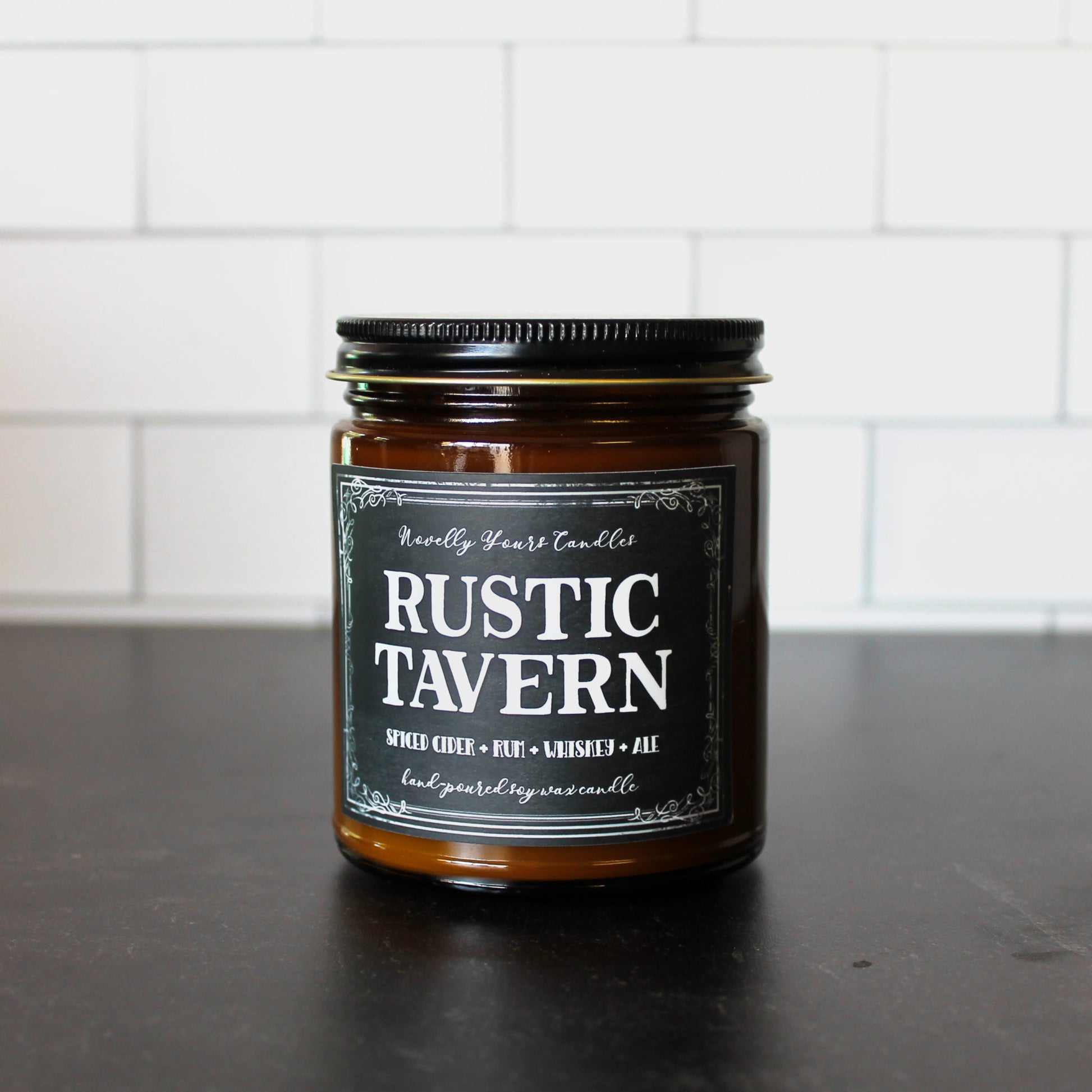 rustic tavern scented soy wax candle in amber jar with black label and black lid on black surface with white tile background