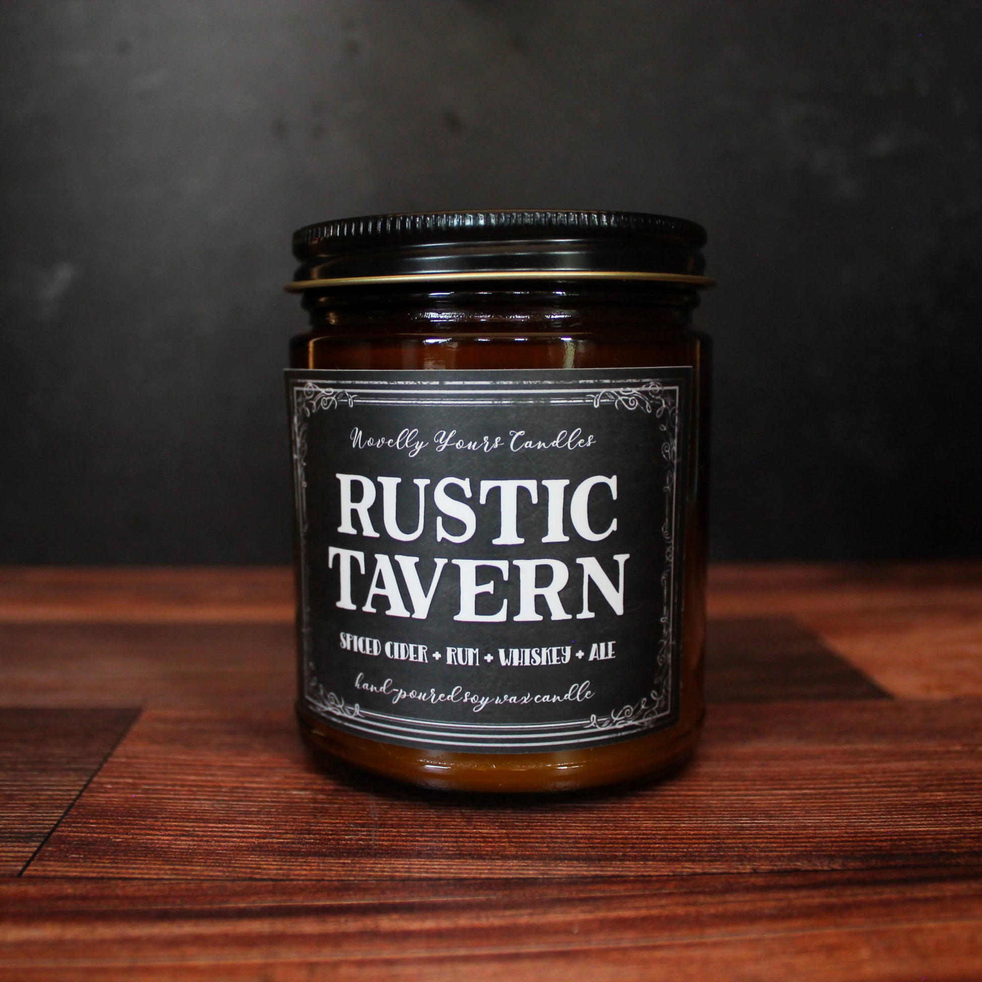 rustic tavern scented soy wax candle in amber jar with black label and black lid on wooden surface with black background