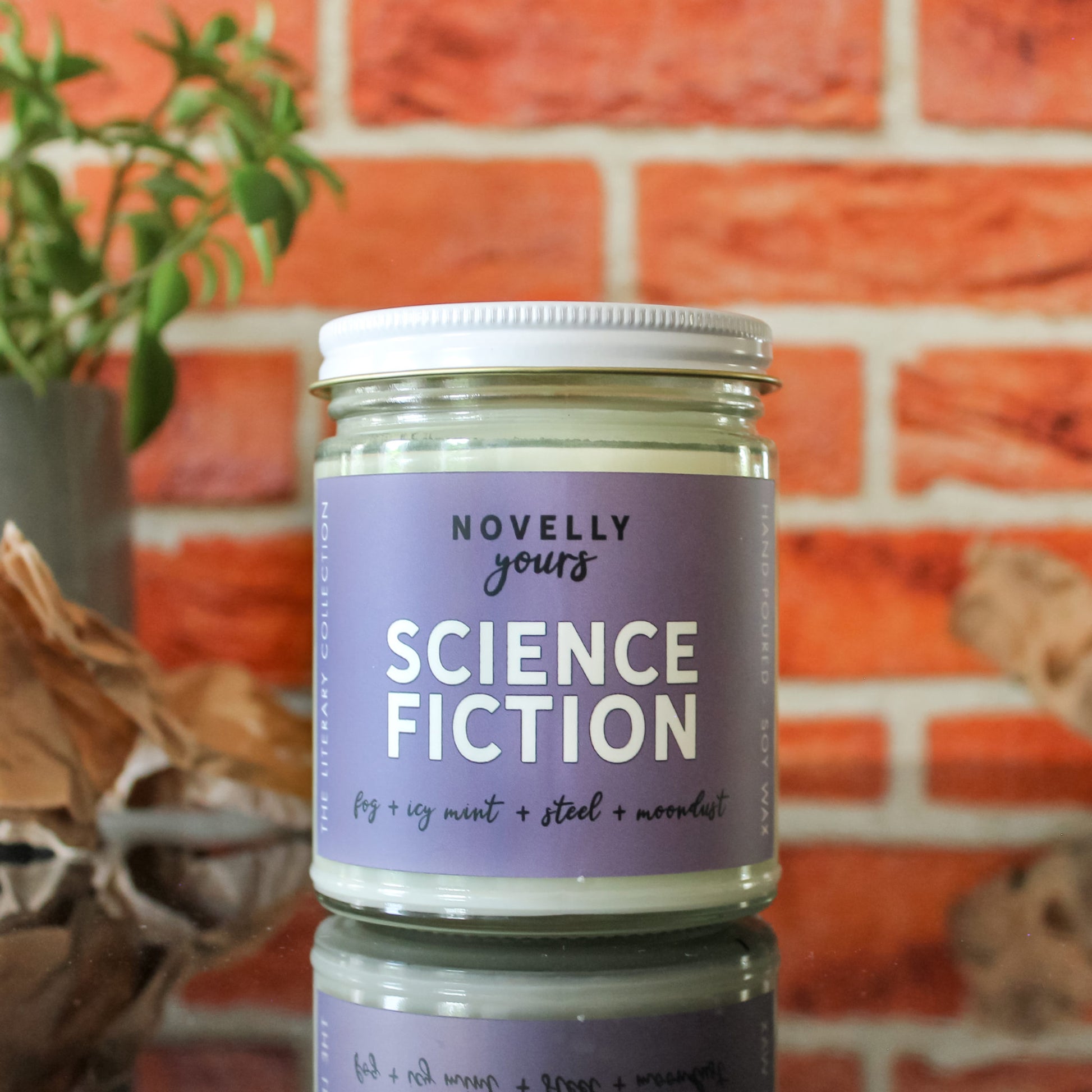 scented soy wax candle labeled "Science Fiction" sitting against a red brick background on a black reflective base