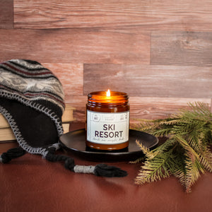 ski resort scented soy wax candle in amber jar, lit surrounded by pine and winter elements