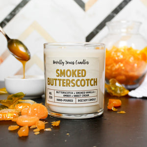 smoked butterscotch scented candle with butterscotch colored text. candle is in clear glass tumbler jar with lid off. candle sits on black surface and is surrounded by butterscotch candy. Spoon full of butterscotch sauce drips in the background