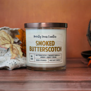 smoked butterscotch scented candle with butterscotch colored text. candle is in clear glass tumbler jar with flat bronze lid. Candle sits on wooden surface with orange background and sweater, fall leaves to accent