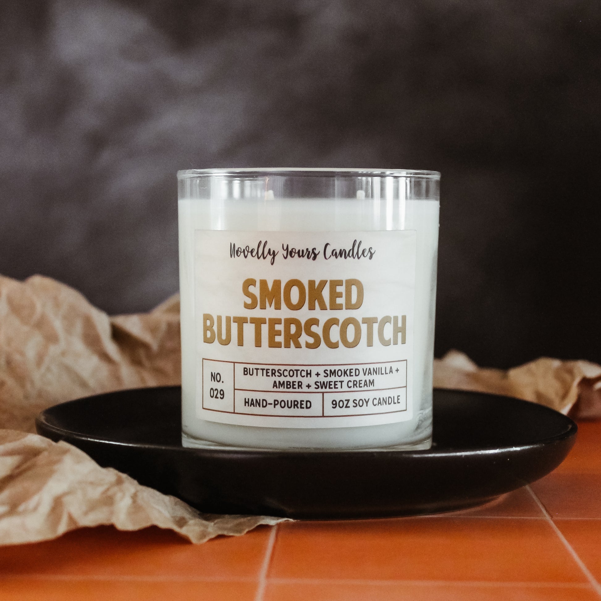 smoked butterscotch scented candle with butterscotch colored text. candle is in clear glass tumbler jar with lid off. Candle sits on round black plate which is on top of orange tiled countertop with smoky black background. brown Kraft paper accents the scene.